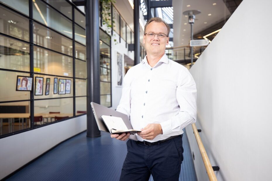 Silmälasipäinen mies seisoo avarassa sisätilassa, vieressä lasiseinää. Käsissä papereita tai vastaavaa.